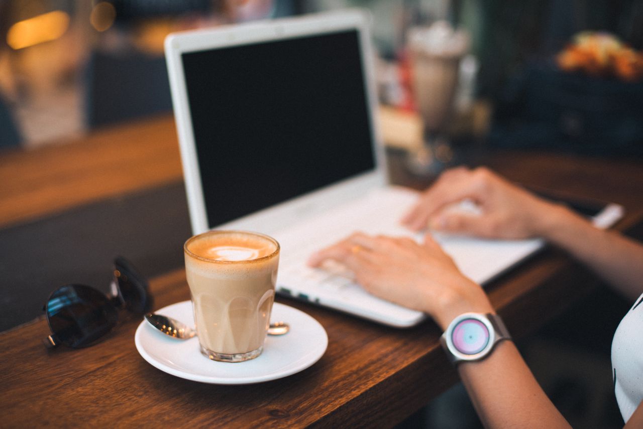 A person typing on a laptop with a latte on a wooden table, indoors.